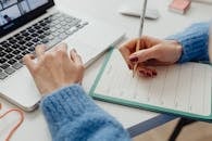A person writes in a planner while using a laptop, focusing on time management.