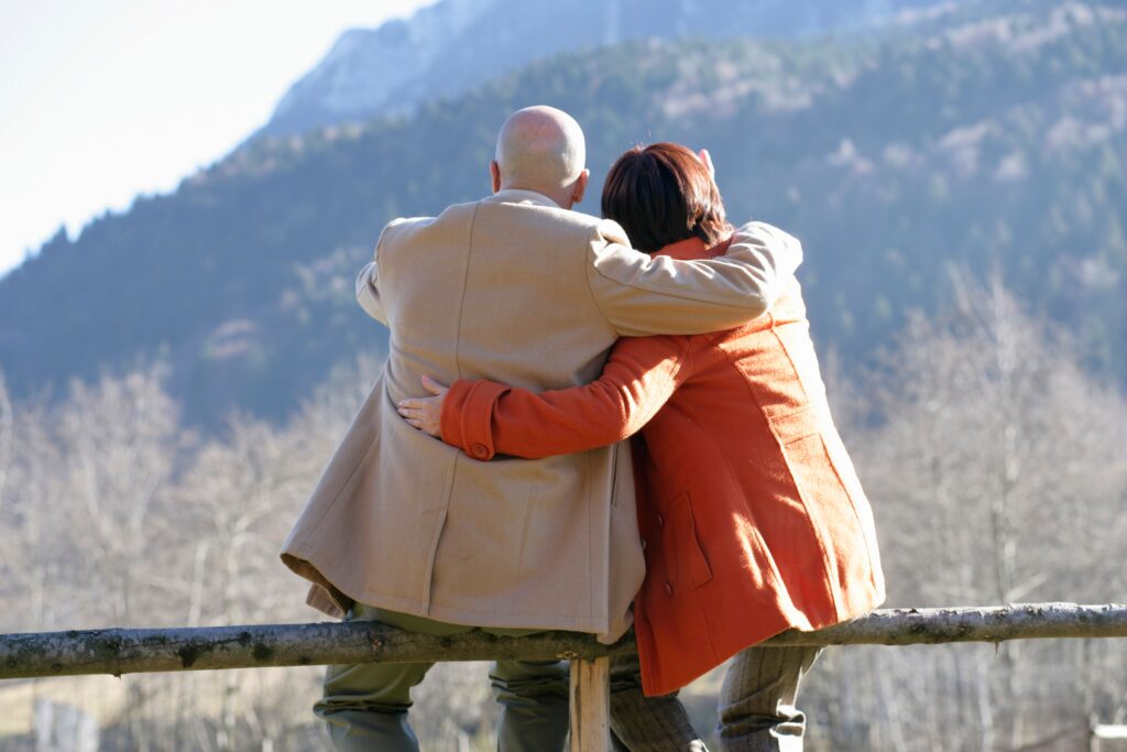 An elderly couple sits together, embracing and enjoying a serene mountain view outdoors.